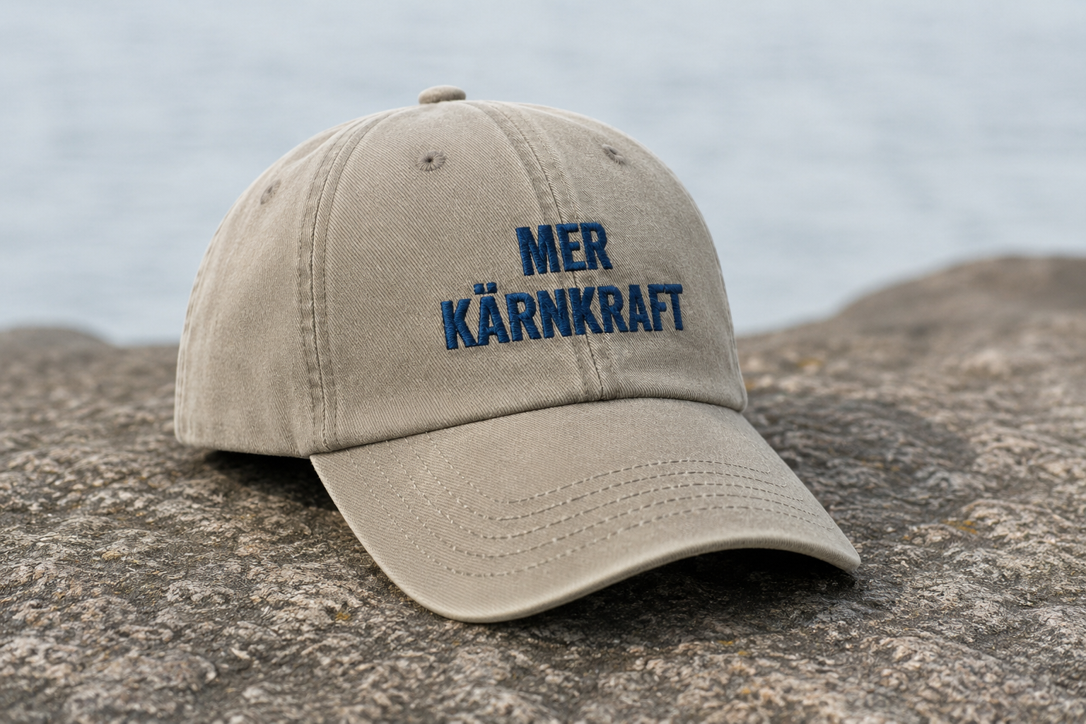 Beige Mer kärnkraft-keps med blå brodyr i skärgårdsmiljö.